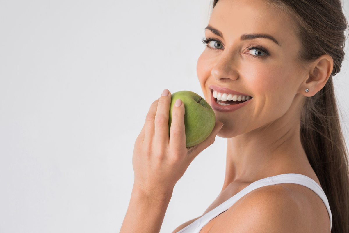 Woman smiles while holding a green apple to her bright, white teeth.